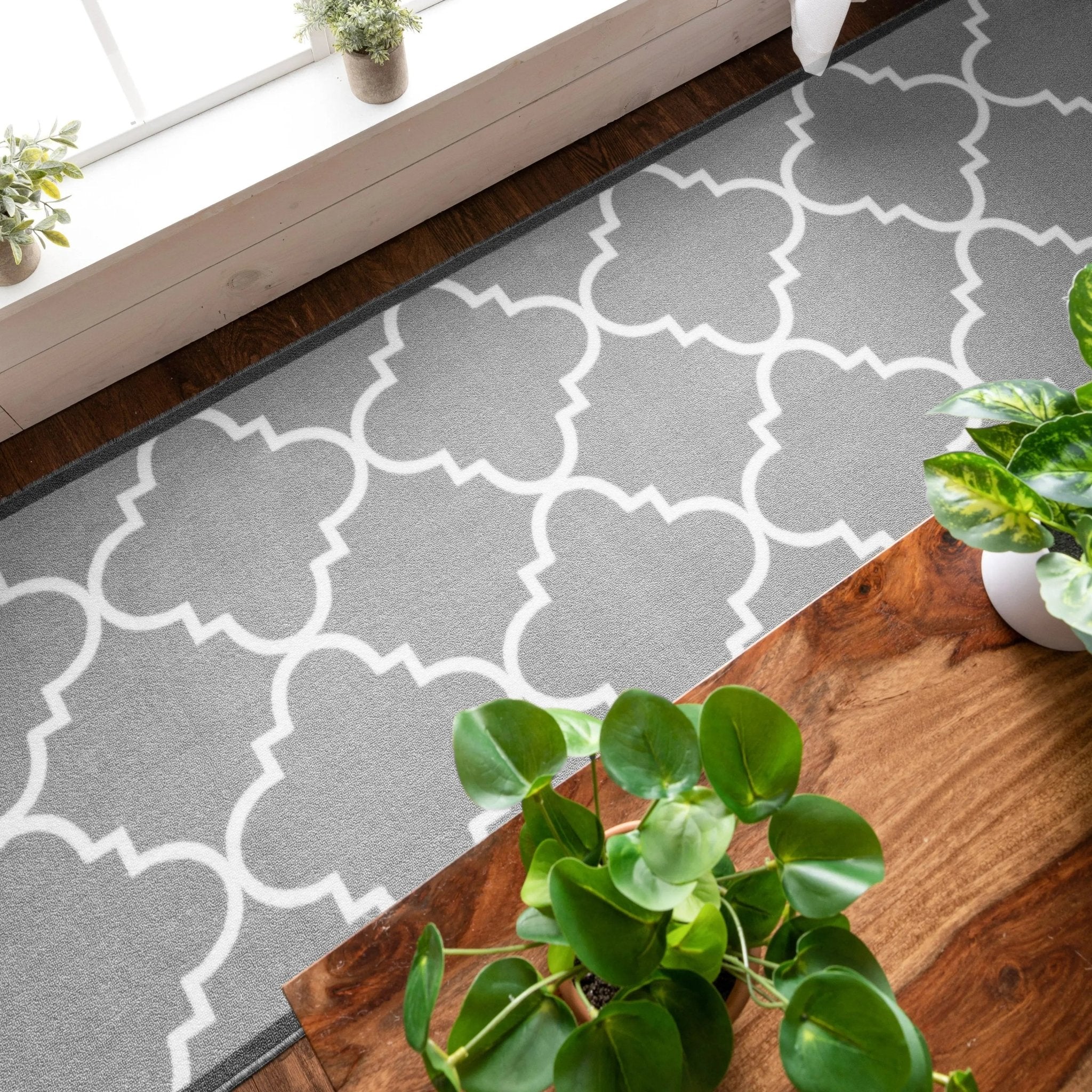 An indoor geometric runner rug shown from above next to a table with plants. It's grey and has a white lattice pattern.